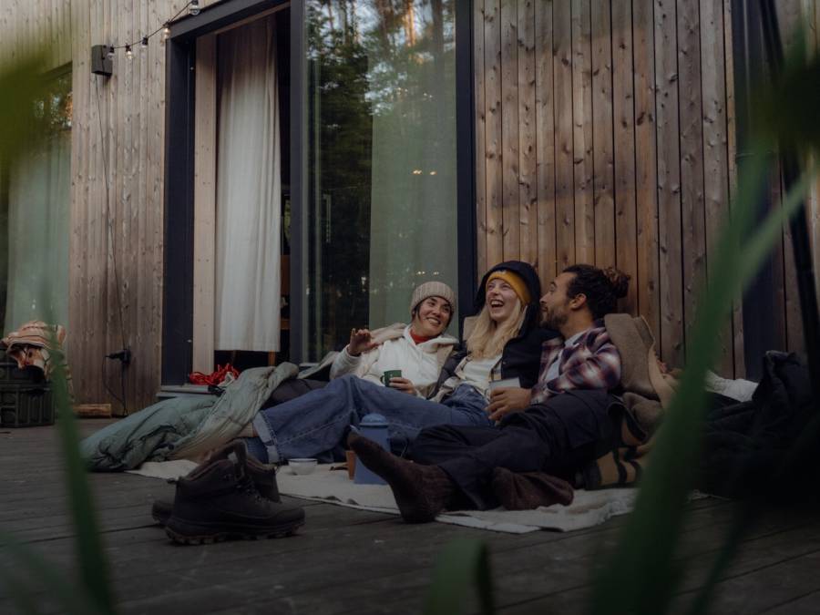 Drie mensen in casual outdoorkleding zitten gezellig bij elkaar op de veranda van een houten hut in het bos