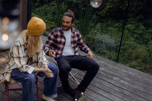 Twee jonge volwassenen in casual outdoorkleding zitten op stoelen voor een houten hut in het bos