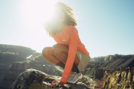 Vrouw met krullend haar en een oranje T-shirt hurkt op een rots vóór een bergachtig landschap.